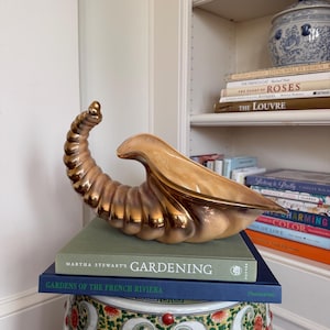 May include: A decorative, gold-toned horn-shaped sculpture rests atop a stack of books. The books include "Gardening" by Martha Stewart and "Gardens of the French Riviera." The sculpture is placed on a patterned garden stool, with a bookshelf in the background.
