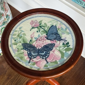 Op de afbeelding: Kleine, ronde houten bijzettafel met een naaldkunst bovenkant. De naaldkunst toont twee donkerblauwe vlinders en roze rozen op een cr&egrave;mekleurige achtergrond, omlijst door een donkerbruine houten rand. De tafel heeft drie gebogen poten.