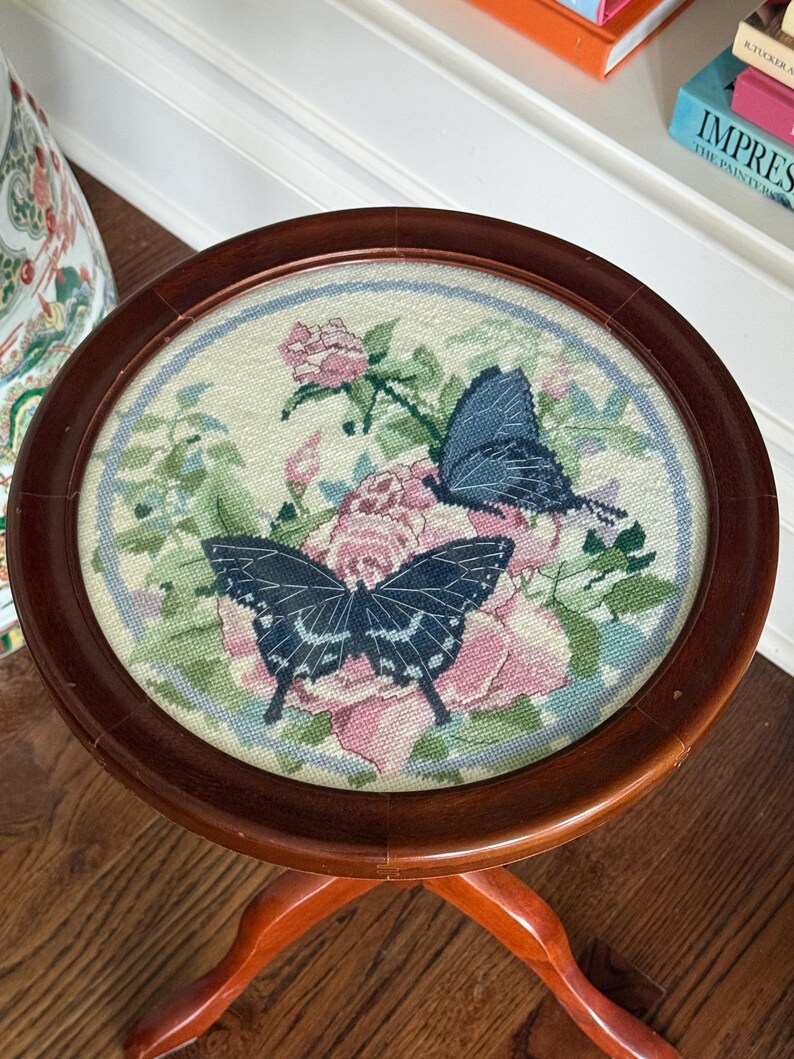 Op de afbeelding: Kleine, ronde houten bijzettafel met een naaldkunst bovenkant. De naaldkunst toont twee donkerblauwe vlinders en roze rozen op een cr&egrave;mekleurige achtergrond, omlijst door een donkerbruine houten rand. De tafel heeft drie gebogen poten.