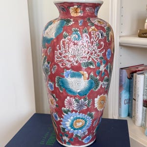 May include: A tall, red ceramic vase with a white rim and a floral design. The vase features large, detailed flowers in shades of pink, blue, and yellow. The vase is sitting on a blue book.