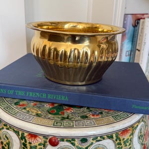 May include: A shiny, gold-colored metal bowl with a wide rim and a ribbed design on the lower half. The bowl is sitting on a dark blue book titled "Gems of the French Riviera". The bowl is a decorative item.