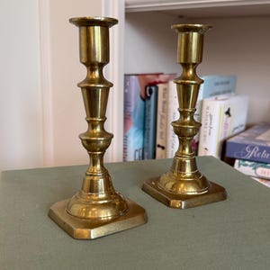 Puede incluir: Dos candeleros antiguos de lat&oacute;n dorado, con base cuadrada y dise&ntilde;o detallado en niveles. Los candeleros son de color oro c&aacute;lido y est&aacute;n sobre una superficie verde. Se ven libros en el fondo.