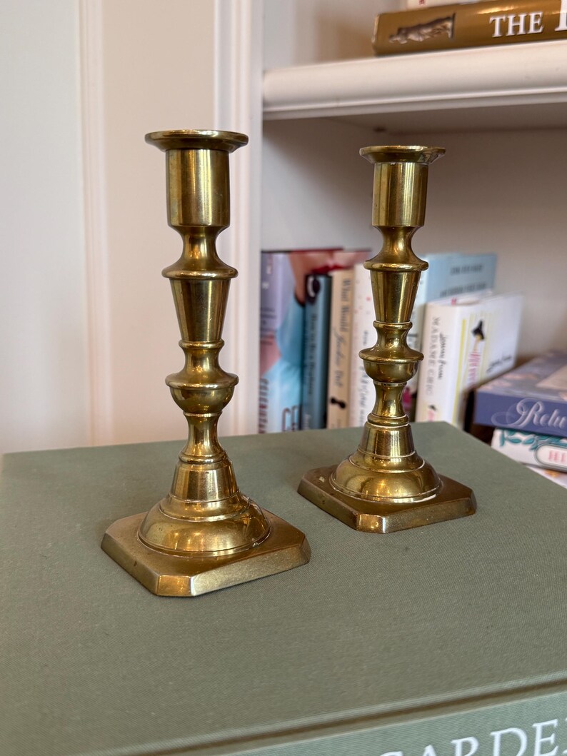 Puede incluir: Dos candeleros antiguos de lat&oacute;n dorado, con base cuadrada y dise&ntilde;o detallado en niveles. Los candeleros son de color oro c&aacute;lido y est&aacute;n sobre una superficie verde. Se ven libros en el fondo.