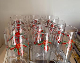 Puede incluir: Un conjunto de vasos transparentes con un diseño festivo de lazo navideño en rojo, verde y dorado. Los vasos están colocados en una estantería con libros, creando un ambiente navideño. Los vasos son cilíndricos y parecen ser de vidrio.