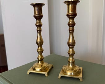 Puede incluir: Dos candelabros antiguos de latón dorado, con bases cuadradas y detalles ornamentados. Los candelabros tienen un tono dorado cálido y están colocados sobre un libro verde. El diseño presenta una parte superior acampanada y un tallo decorativo.
