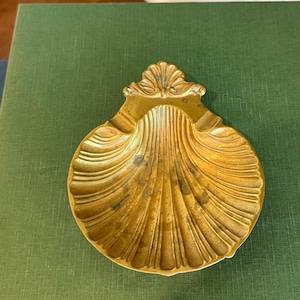 May include: A gold-colored, seashell-shaped dish with a detailed, ridged design. The dish has a decorative, ornate top and is resting on a green surface. The dish is likely made of metal and is a decorative item.