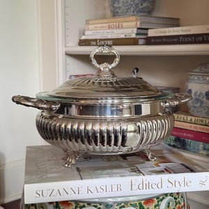 May include: A silver-plated serving dish with a matching lid, featuring a fluted body, two ornate side handles, and four decorative feet. The lid is topped with a central handle and the dish rests on a book titled Suzanne Kasler Edited Style.