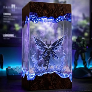 May include: A rectangular lamp featuring dark wood top and bottom sections with a clear resin centre. Inside the resin, a dark, winged humanoid figure is surrounded by glowing blue and purple light effects that resemble swirling energy or smoke.