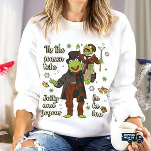 Puede incluir: Sudadera blanca con un diseño navideño con Kermit la Rana y otro personaje de rana. El diseño incluye el texto "Tis the season to be Jolly and joyous, la la laa". Las ranas visten ropa de invierno.