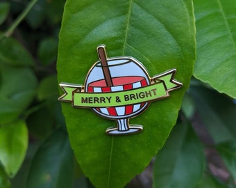 May include: A festive enamel pin featuring a cocktail design with a red and white striped drink, a brown straw, and a green banner that reads "MERRY & BRIGHT". The pin is set against a green leaf background.