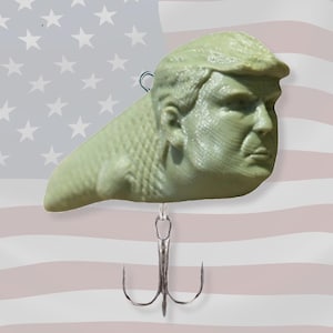 May include: A light green fishing lure shaped like a head with a prominent hairstyle. The lure has a silver hook with three barbs. The background features a waving American flag.