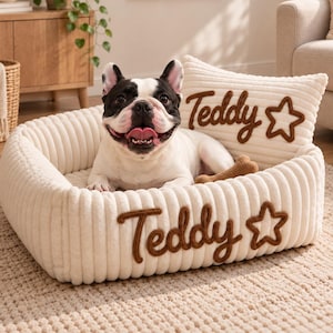 Puede incluir: Una cama para mascotas de color crema con textura acanalada y un cojín trasero a juego. La parte frontal de la cama y el cojín presentan el nombre Teddy bordado en letra cursiva marrón, junto con el contorno de una estrella marrón.
