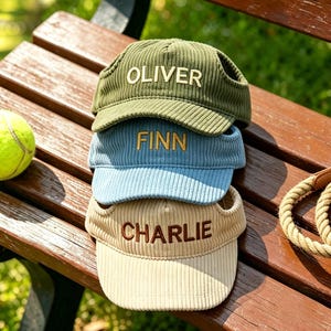 May include: Three corduroy dog visors stacked on a wooden bench. The visors are olive green, light blue, and beige, each embroidered with a name in a matching color. A tennis ball and a dog leash are also on the bench.