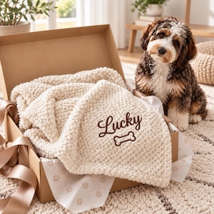 Pode incluir: Uma manta de malha texturizada na cor creme dobrada dentro de uma caixa de presente de papelão. A manta apresenta o texto bordado em marrom Lucky acima de um ícone de osso marrom. Um cão marrom, branco e preto senta-se ao lado da caixa.
