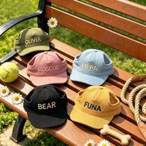May include: Five corduroy dog hats in olive green, pink, light blue, black, and yellow. Each hat has ear holes and a name embroidered on the front: OLIVER, ROSCOE, PIPPA, BEAR, and FUNA. A tennis ball and daisy flowers are also visible.