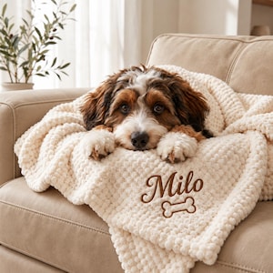 Pode incluir: Uma manta de malha texturizada na cor creme, com o nome Milo bordado em letras cursivas castanhas acima de um ícone de osso de cão castanho. Um cachorro castanho, branco e preto descansa a cabeça sobre a manta dobrada.