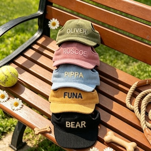 May include: A stack of corduroy dog hats in olive green, pink, light blue, yellow, and black, each embroidered with a name in a matching color. The hats are on a wooden bench with a tennis ball and daisies.