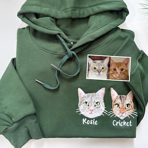 Peut inclure: Sweat à capuche vert foncé avec cordon de serrage et poignets côtelés. Comprend une petite photo de deux chats et des visages de chats brodés avec les noms "Rosie" et "Cricket" en dessous. Vêtement décontracté et confortable.