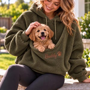 Op de afbeelding: Een olijfgroene fleece hoodie met een grote voorzak ontworpen voor het dragen van een klein huisdier. De hoodie heeft een capuchon met trekkoord, lange mouwen met geribbelde boorden en de naam Buddy in bruin geborduurd met een pootafdruk.