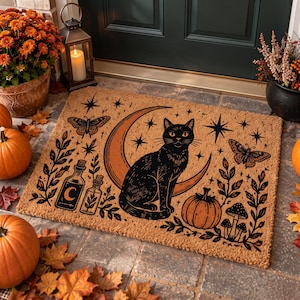 May include: A rectangular tan coir doormat featuring a black ink illustration of a cat sitting in front of a crescent moon. The design includes butterflies, stars, two glass bottles, a pumpkin, mushrooms, and leafy branches in a dark print.