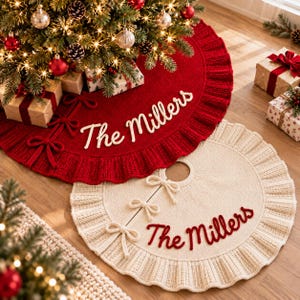 Puede incluir: Una falda de árbol de Navidad circular de punto rojo con borde con volantes, tres cierres de lazo y el texto bordado en blanco "The Millers", junto a una falda de árbol de punto color crema a juego con el texto bordado en rojo "The Millers".