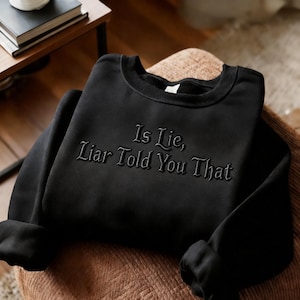 Può includere: Felpa nera con la scritta ricamata "Is Lie, Liar Told You That". La felpa girocollo è piegata su una superficie intrecciata marrone. Le maniche sono arrotolate.