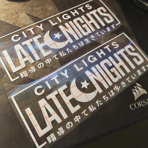 Könnte beinhalten: Zwei rechteckige Aufkleber mit dem weißen Text CITY LIGHTS LATE NIGHTS und einem Halbmond- und Sternsymbol. Unter dem englischen Text sind japanische Schriftzeichen gedruckt. Die Aufkleber haben eine mit einem Raster versehene Transferfolie.