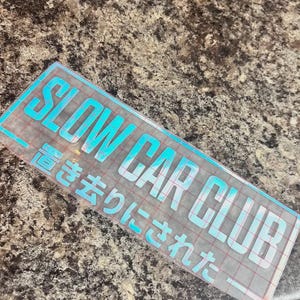 Könnte beinhalten: Ein rechteckiger Vinyl-Aufkleber mit hellblauer, irisierender Oberfläche, der den Text SLOW CAR CLUB in einer fetten serifenlosen Schrift zeigt, gefolgt von dem japanischen Text 置き去りにされた, alles auf einem transparenten Transferband mit rotem Gittermuster.