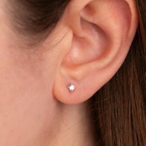 May include: A close-up of an ear with a small, square stud earring. The earring is a light colour, possibly silver or white gold, and has a single, clear stone. The person has long, brown hair.