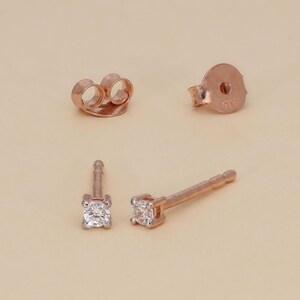 May include: Rose gold stud earrings with a square, clear gemstone. The earrings are shown with two butterfly backs and a single post back. The earrings are displayed on a light beige background.