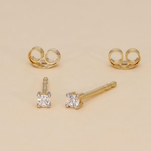 May include: A pair of gold stud earrings with square, clear gemstones. Two gold earring backs are also visible. The earrings are displayed on a light beige surface.
