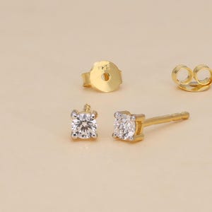 May include: A pair of gold stud earrings with a square, clear gemstone setting. The image also shows two earring backs. The earrings are displayed on a light beige surface.