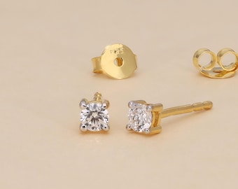 May include: A pair of gold stud earrings with a square, clear gemstone setting. The image also shows two earring backs. The earrings are displayed on a light beige surface.