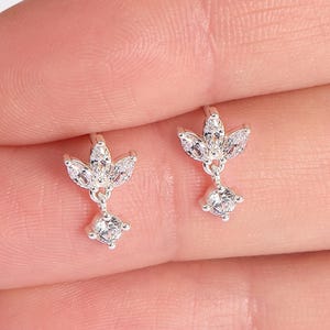 May include: A pair of silver earrings. Each earring has a floral design at the top with a dangling square gemstone. The earrings are held in a person's hand.