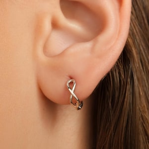 Peut inclure: Gros plan d'une boucle d'oreille argentée en forme de symbole infini sur une oreille. La boucle d'oreille est un petit cercle délicat avec un design entrelacé. La peau est claire et les cheveux sont bruns foncés.
