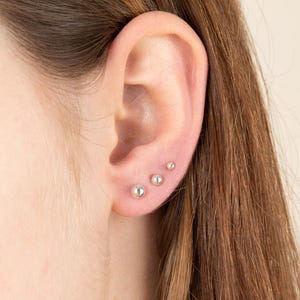 May include: An ear with three silver stud earrings of varying sizes. The earrings are small, round, and positioned in a line. The largest earring is at the bottom, with the size decreasing upwards. The background is a neutral colour.