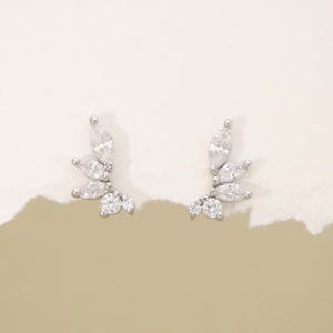 May include: A pair of silver ear climbers, each with a cluster of clear marquise-cut and round gemstones. The earrings are arranged in a leaf-like design, creating a delicate and elegant appearance. The background is a soft, neutral colour.