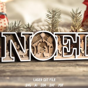 May include: A wooden Christmas decoration spelling "NOEL" with a layered design. The center of the "O" features a nativity scene. The letters are white with a dark brown outline. The background includes Christmas decorations and greenery.