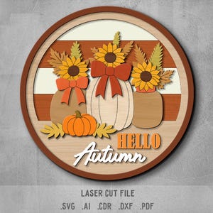 May include: A circular wooden sign with an autumnal theme. It displays three pumpkins adorned with sunflowers and bows, a small orange pumpkin, and the words "HELLO Autumn". The sign is a laser cut file.