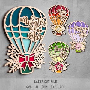 May include: Wooden laser-cut hot air balloon decorations, each representing a season: Winter, Autumn, Spring, and Summer. The Winter balloon is teal, the Autumn balloon is orange, the Spring balloon is purple, and the Summer balloon is green. Each balloon has the season's name in script.