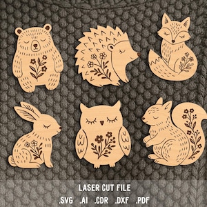 May include: Wooden laser-cut animal shapes, including a bear, hedgehog, fox, rabbit, owl, and squirrel. Each animal has a floral design and closed eyes. The text "LASER CUT FILE" and file type extensions are at the bottom.