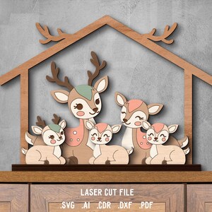 May include: Wooden laser-cut family of deer, featuring five deer figures in various sizes, with brown antlers, and pink and green accents. The deer are arranged under a wooden house-shaped frame. The text "LASER CUT FILE" is at the bottom.