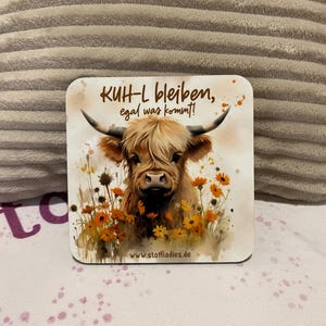 Puede incluir: Posavasos cuadrado con una ilustración de acuarela de una vaca de las Tierras Altas con largo pelaje marrón y desgreñado, rodeada de flores naranjas. El posavasos tiene el texto "KUH-L bleiben, egal was kommt!" y el sitio web www.stoffladies.de.