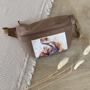 Può includere: Un marsupio marrone con un dipinto ad acquerello di un serpente. La borsa ha uno scomparto principale con cerniera e una tracolla regolabile. Lo sfondo è un tessuto a nido d'ape beige chiaro e una superficie in legno.