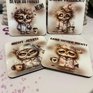May include: Set of four square coasters featuring sleepy owl illustrations in pajamas, each holding a coffee cup. Text on the coasters includes phrases like "NO!" and "NOT NOW!" in German. The coasters have a white background.