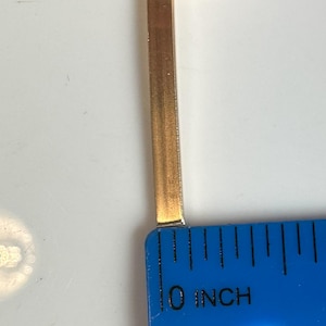 May include: A gold-coloured metal bar is positioned next to a blue ruler. The ruler shows the bar's length, starting at the 0-inch mark. The bar appears to be approximately 0.64 cm long.