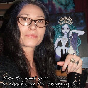 May include: A woman with long dark hair and glasses is holding a print of a blue-haired mermaid with a crown and a lantern. The print has a starry background and the text "MYKA ELUNA" is at the bottom. The woman is smiling and pointing to the print.
