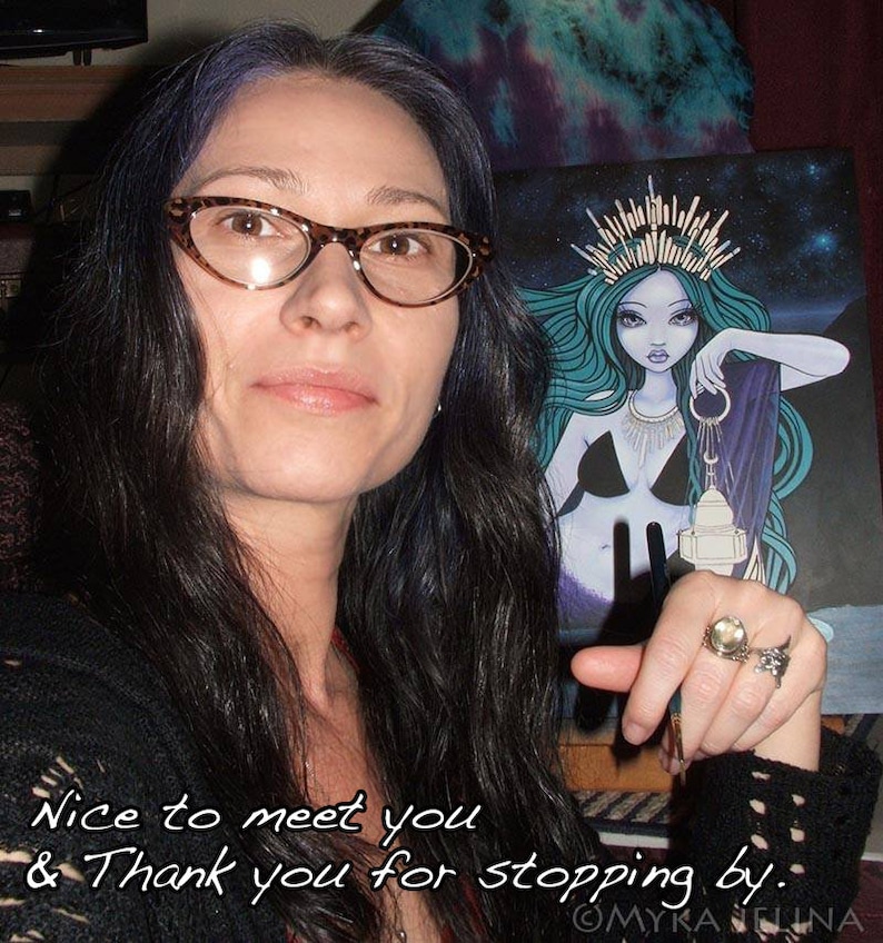 May include: A woman with long dark hair and glasses is holding a print of a blue-haired mermaid with a crown and a lantern. The print has a starry background and the text "MYKA ELUNA" is at the bottom. The woman is smiling and pointing to the print.