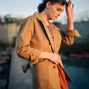 Op de afbeelding: Een beige blazer met een dubbele rij knopen, manchetten met knopen en een ceintuur. De blazer wordt gecombineerd met een roestkleurige rok. De persoon draagt een zonnebril.