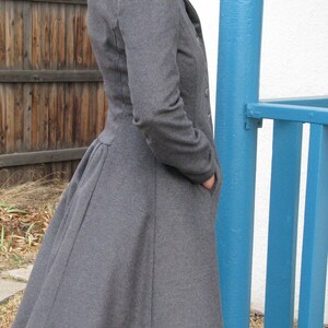 May include: A gray, long coat with a flared skirt and a tailored fit. The coat has a classic design with a notched collar and button closures. The model is wearing the coat with their hands in the pockets. The coat is made of a soft, wool-like material.